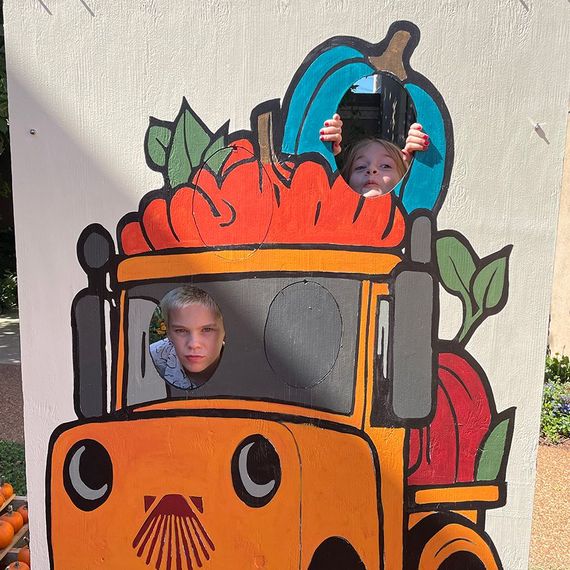 Two children pose in a pumpkin truck cutout, one in the cab, another in the pumpkin bed.