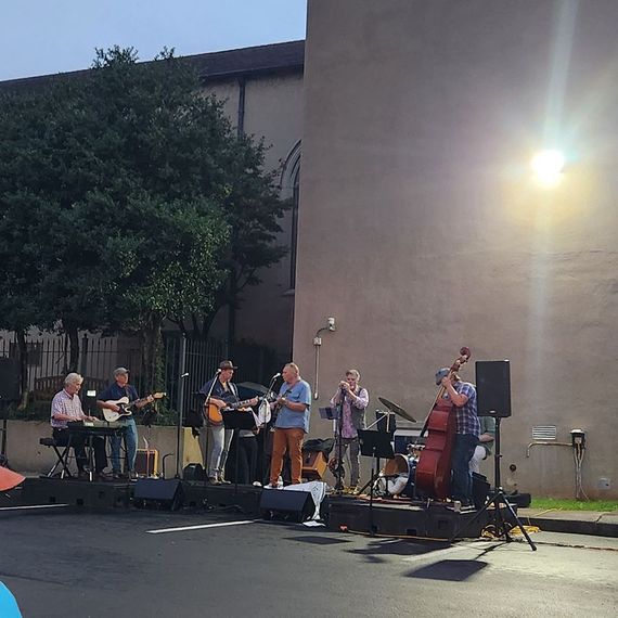 A band performing outdoors. Musicians playing instruments in front of a building, under a light, at dusk.