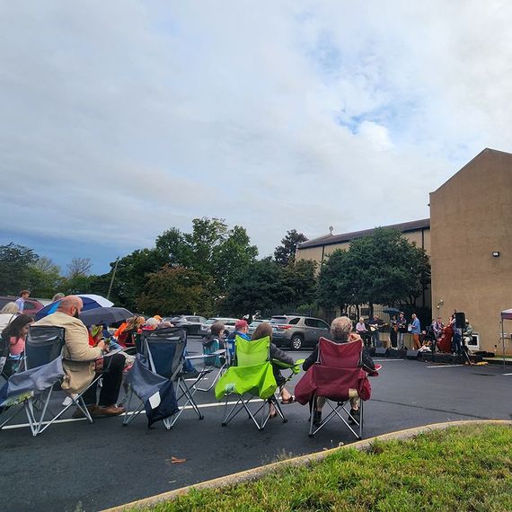 People in folding chairs at an outdoor event in front of a building, under a cloudy sky.