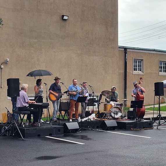 A band performs outdoors: musicians on stage playing instruments, under overcast skies.