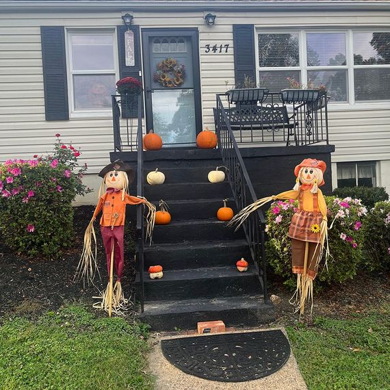Two scarecrows flank a porch decorated with pumpkins. Door has a wreath. House is light colored.