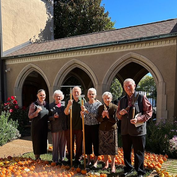 Group of seniors holding pumpkins, standing in front of arched building, surrounded by pumpkins.