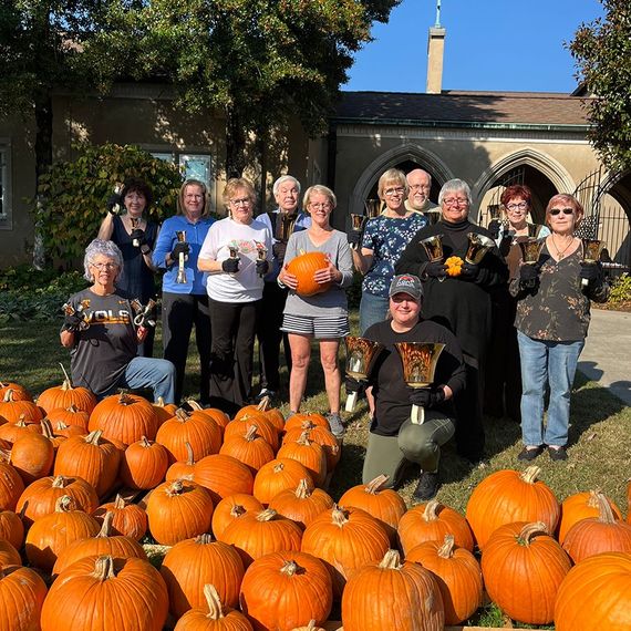 Group of people holding bells and pumpkins in front of a building. Sunny, outdoor setting.