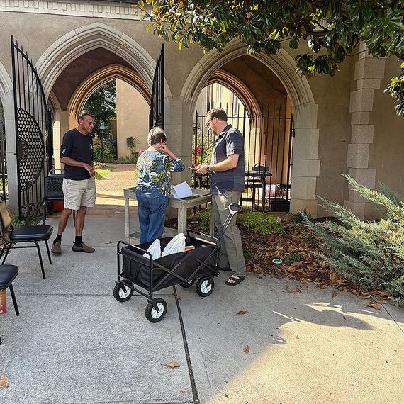 Three people at a table outside a building with arched entryways. A wagon sits in front of the table.