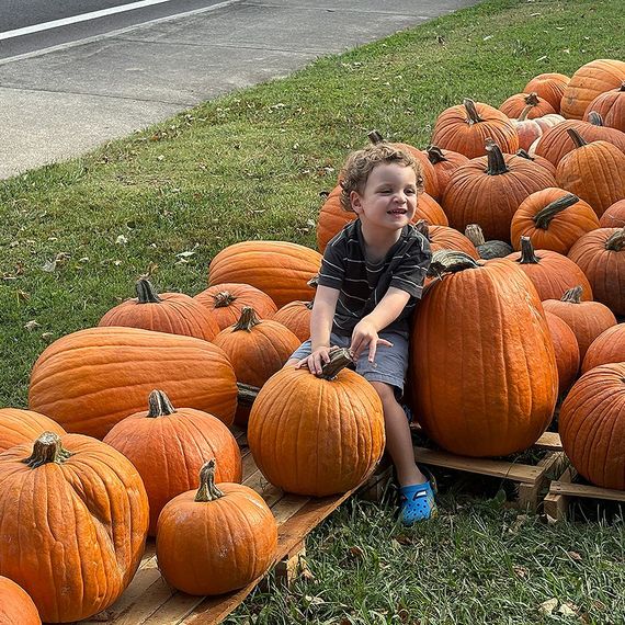 Boy sitting on a pumpkin in a pumpkin patch; outdoors, smiling.