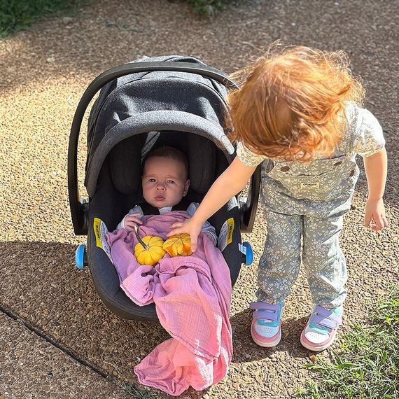 Young child touches baby in a car seat outdoors. The baby is wrapped in a pink blanket and has small pumpkins.