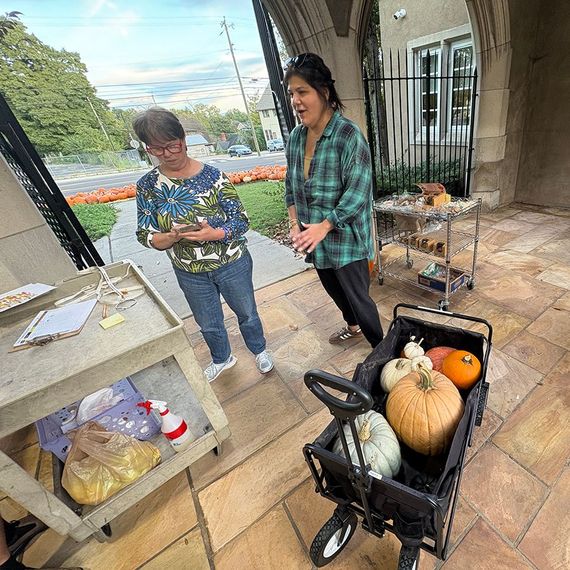 Two women outside, one looking at a phone, the other gesturing excitedly. A wagon of pumpkins sits nearby.