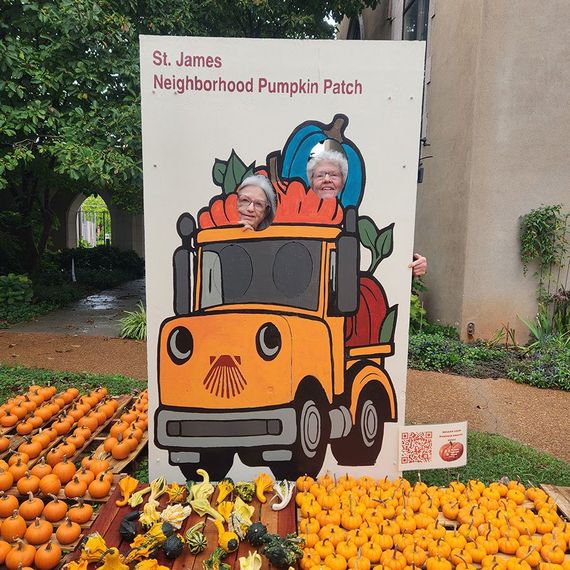 Two women pose behind a cutout of a pumpkin truck at St. James Pumpkin Patch, surrounded by pumpkins.