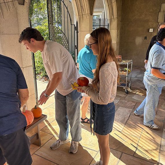 People outdoors at a gathering, including a young woman holding an apple and a young man looking at pumpkins.