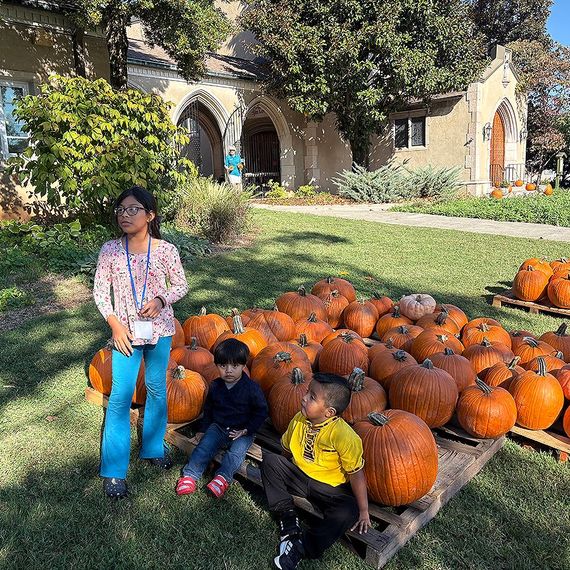 Children pose by pumpkins on a pallet outdoors, church in the background.