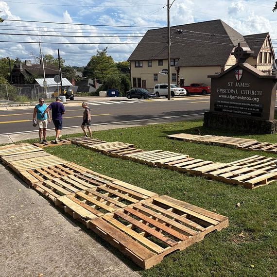 People near wooden pallets laid out on a church lawn, next to a road.