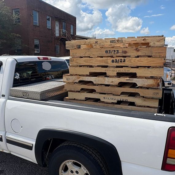 White pickup truck bed loaded with wooden pallets, parked outside a brick building.
