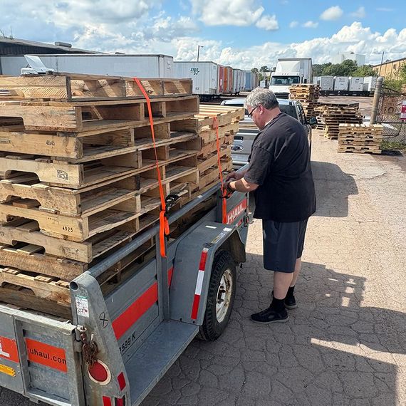 Man straps down a stack of wooden pallets on a trailer. Outdoors, sunny day.