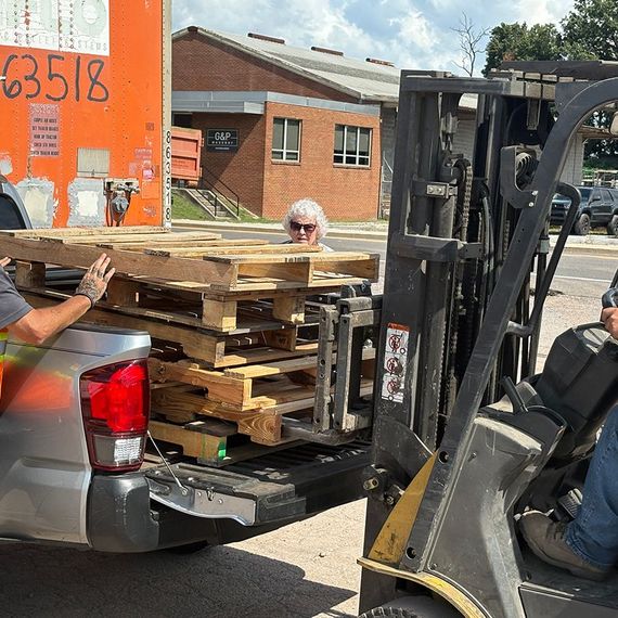 Forklift loading pallets into a pickup truck at an outdoor setting, two people assisting.