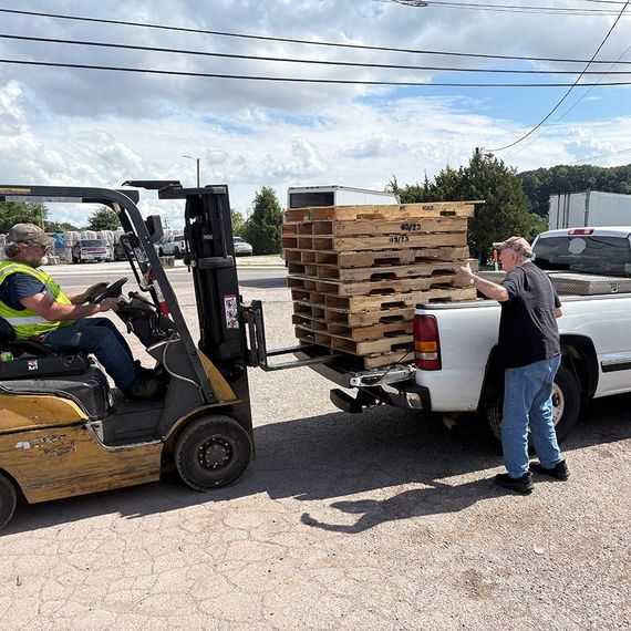 Forklift loading wooden pallets onto a white pickup truck; two men outside near power lines and a building.