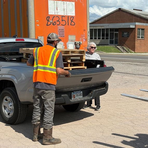 Man in orange vest loads pallets into a truck with a woman watching near an orange container and building.