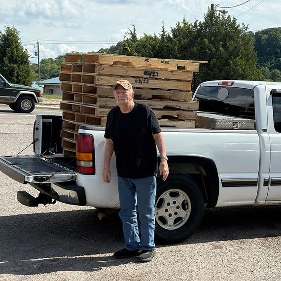Man standing next to white pickup truck loaded with wooden pallets, outdoors.