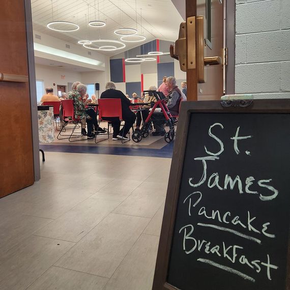 View of a St. James Pancake Breakfast. People seated at tables in a brightly lit room. A chalkboard sign is in the foreground.