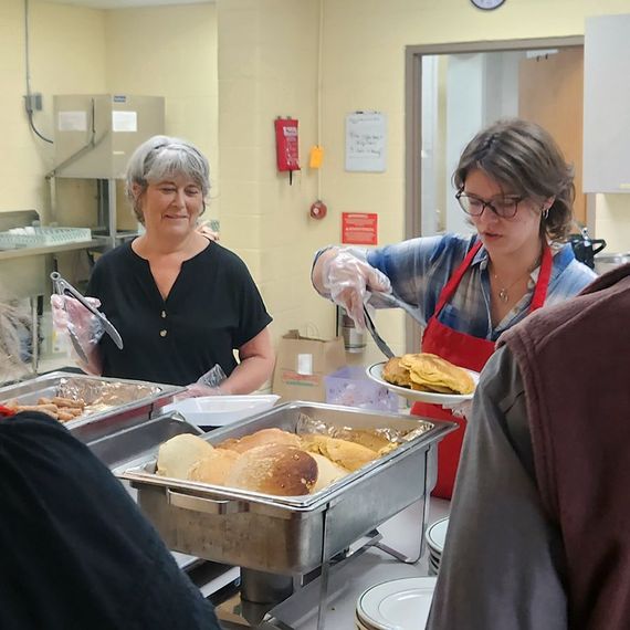 Two people serving food at a buffet. One person is wearing a red apron and serving from a pan.