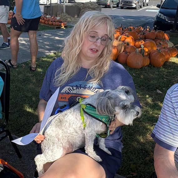 Woman with glasses, holding a small gray dog on her lap. Pumpkins are visible in the background.
