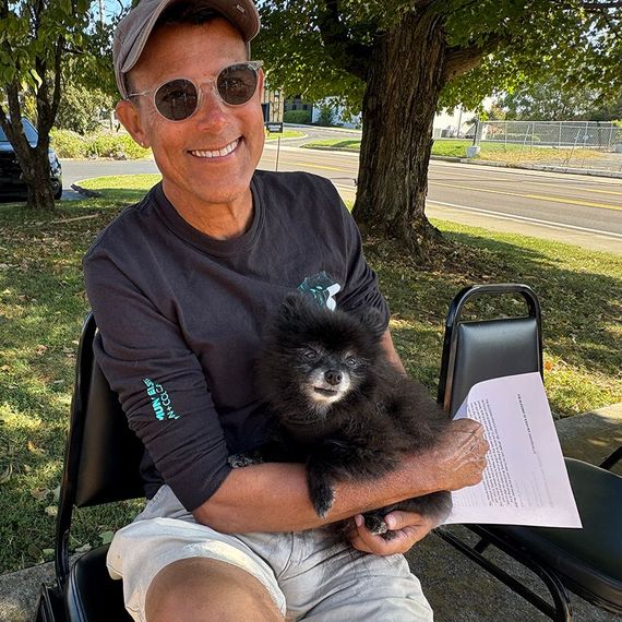 Man smiling, holding black Pomeranian dog. Outdoors, wearing sunglasses, casual clothes.