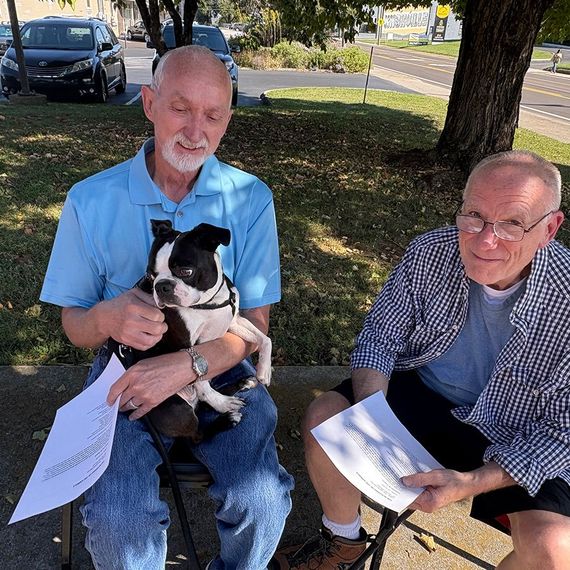 Two men sitting outside, one holding a Boston terrier dog. They hold papers.