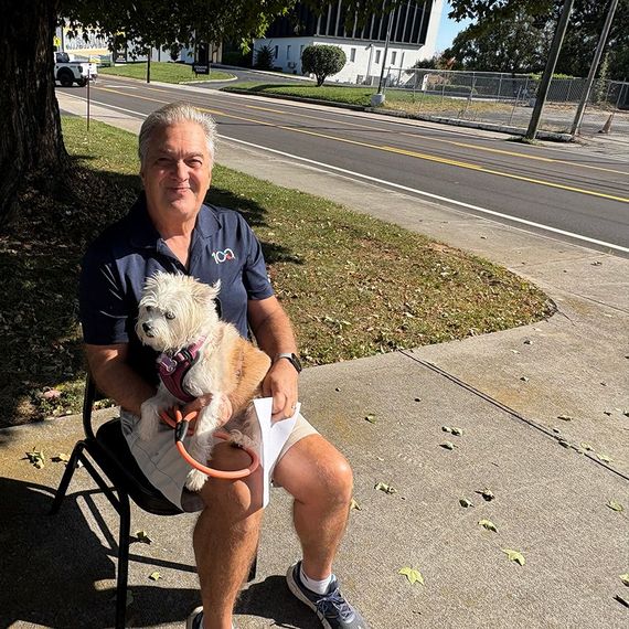 Man in blue shirt holds small tan dog on his lap, sitting outside.