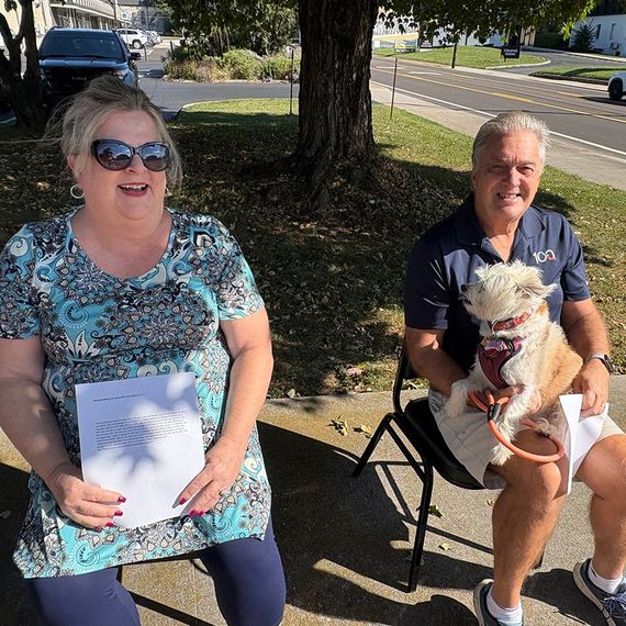 Woman smiles, holds papers. Man with dog sits outdoors, near a road.