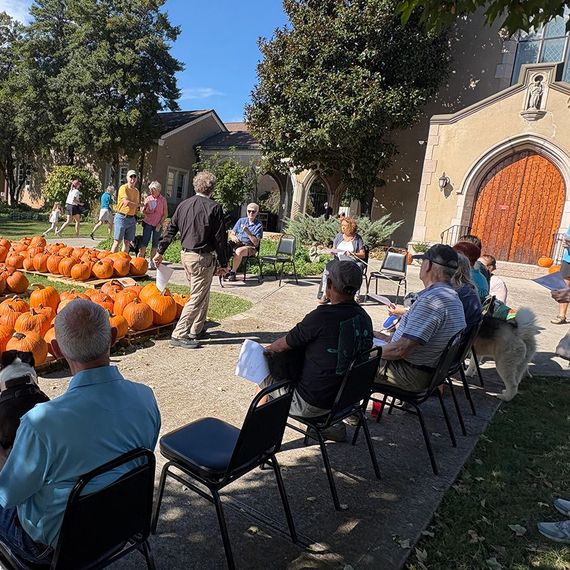 A group of people at an outdoor event. A man speaks near pumpkins, a church entrance is in the background.
