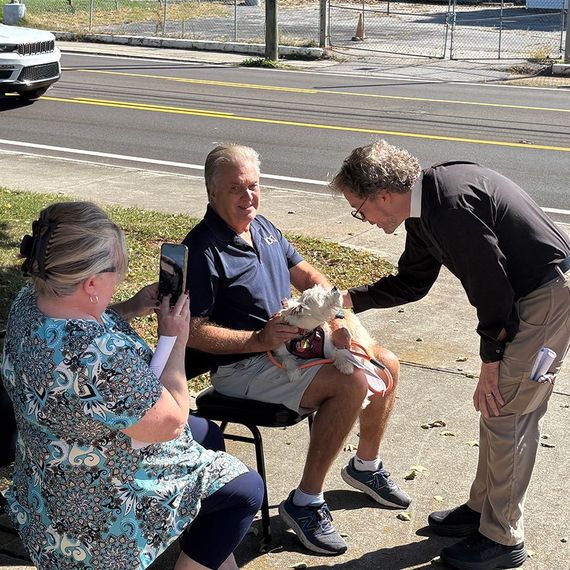 A man pets a small dog held by another man while a woman photographs them outdoors.
