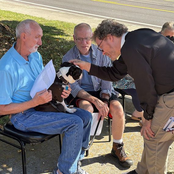 Three men petting a Boston Terrier, outdoors. Man in blue shirt holds dog, another man looks on.