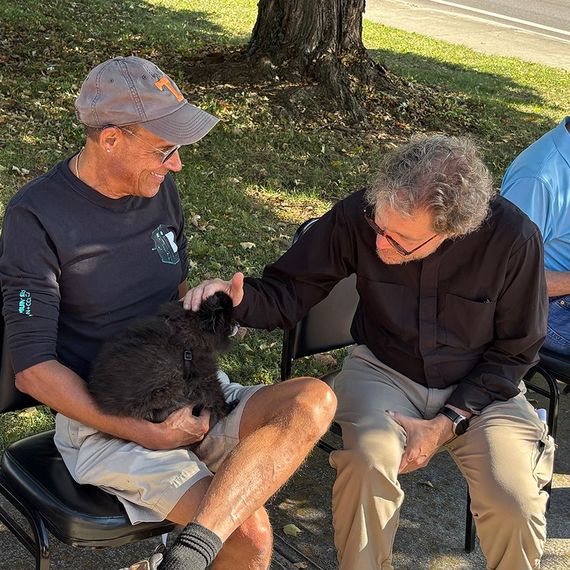 Two men seated outdoors petting a small black cat. One man wears a cap, the other glasses.