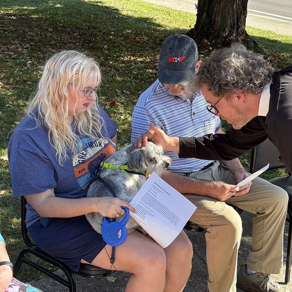 Woman with dog, man petting dog, and man reading paper outdoors.