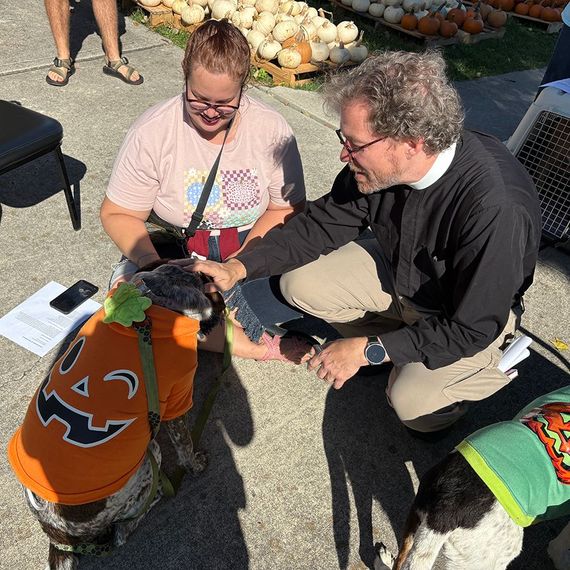 Two people pet dogs in Halloween costumes at an outdoor event with pumpkins in the background.