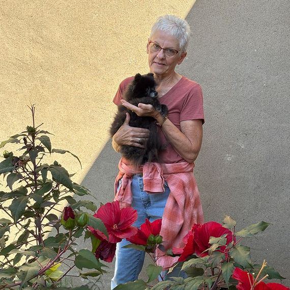Woman with short gray hair holds a black dog in front of a tan wall and red flowers.