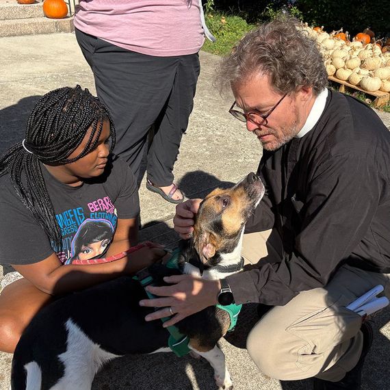 A Black teen and a white man pet a dog. Pumpkins in background.