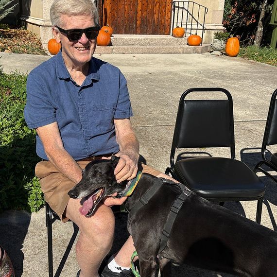 Man in sunglasses petting a black dog outdoors; pumpkins and church doors in the background.
