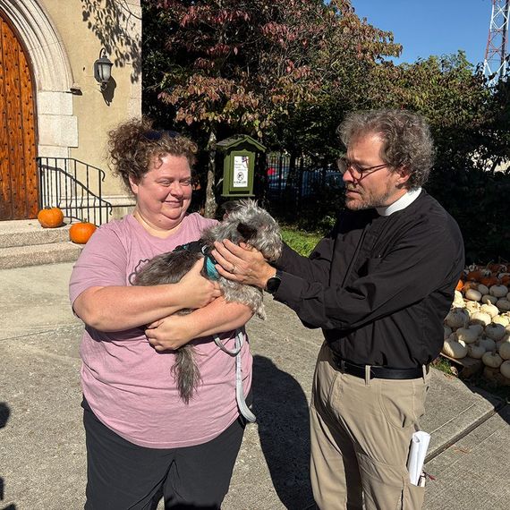 Woman holding dog, being blessed by a priest outside a church with pumpkins.