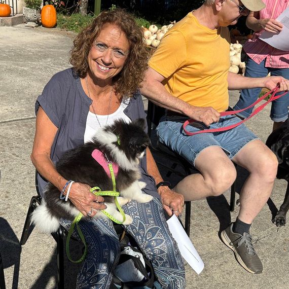 Woman holding a black and white dog outdoors; smiling; man nearby. Pumpkins in background.