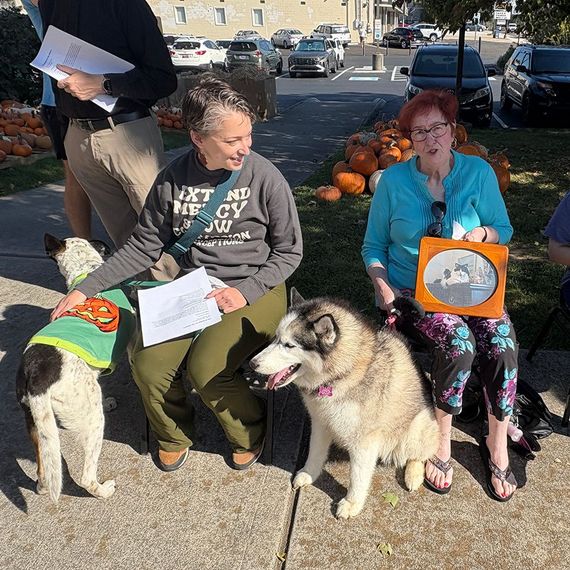 Two women seated with dogs, holding papers and a framed photo outdoors near pumpkins and parked cars.