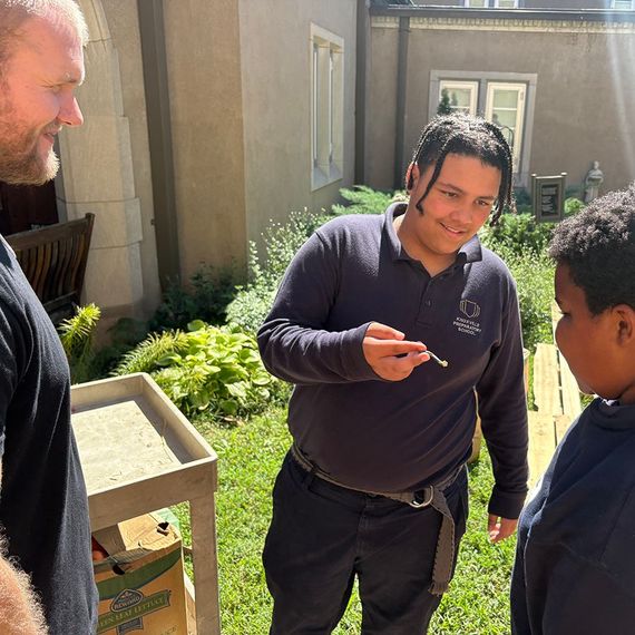 Three people outside: one man, two boys. The man and a boy watch the other boy hold a small object, smiling.