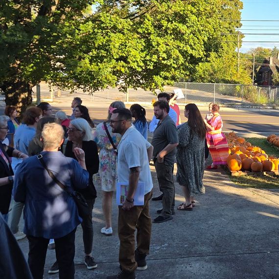 Group of people outside near pumpkins and a building. Sunlight and greenery are visible.