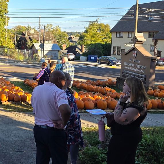 People by a pumpkin patch in front of a church. Some are conversing; others are browsing pumpkins.