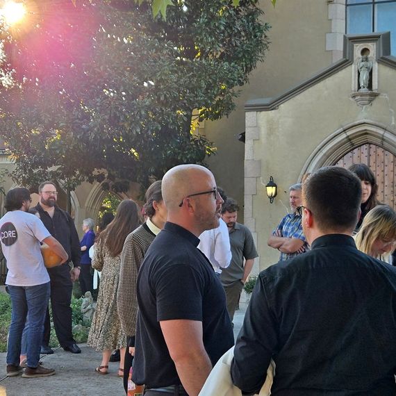People gathered outside a building with gothic architecture; sunlight streams from behind a tree.