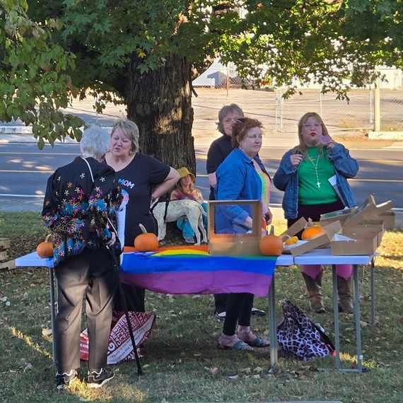Group of people at a table with a LGBTQ+ flag, pumpkins, and items. Outdoors, sunny day.