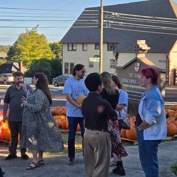 Group of people near pumpkins, in front of church. Sunny, warm tone. Some are talking, others looking on.
