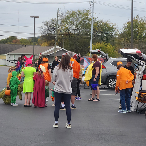 People in costumes at a trunk-or-treat event in a parking lot. Cars with open trunks and decorations.