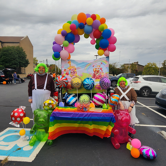 Two people in costume stand by a candy-themed car display, under a balloon arch.