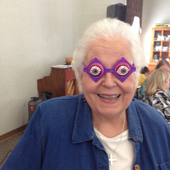 Older woman smiling, wearing purple glasses with eyeballs. Denim shirt, crucifix necklace, piano in background.