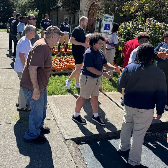 People gathered outdoors near pumpkins. Some reaching toward them. A sign displays a handicap symbol.